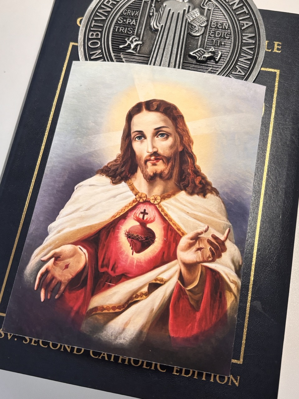 Ein religiöses Bild von Jesus Christus mit einem Herzsymbol, vor einem schwarzen Bucheinband mit goldener Beschriftung. Sacraresponda